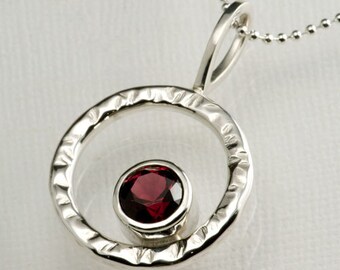 May include: A silver pendant necklace featuring a textured circular design with a red gemstone at its centre.