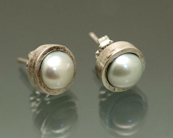 Puede incluir: Un par de pendientes de plata con una perla blanca en cada uno. Los pendientes tienen un engaste de plata texturizado.