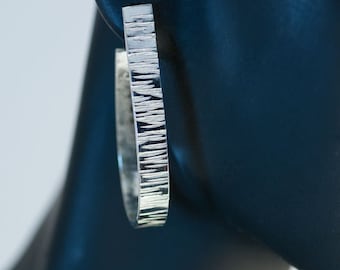 Puede incluir: Pendientes de aro de plata con un acabado texturizado martillado. Los pendientes tienen un diseño simple y moderno.