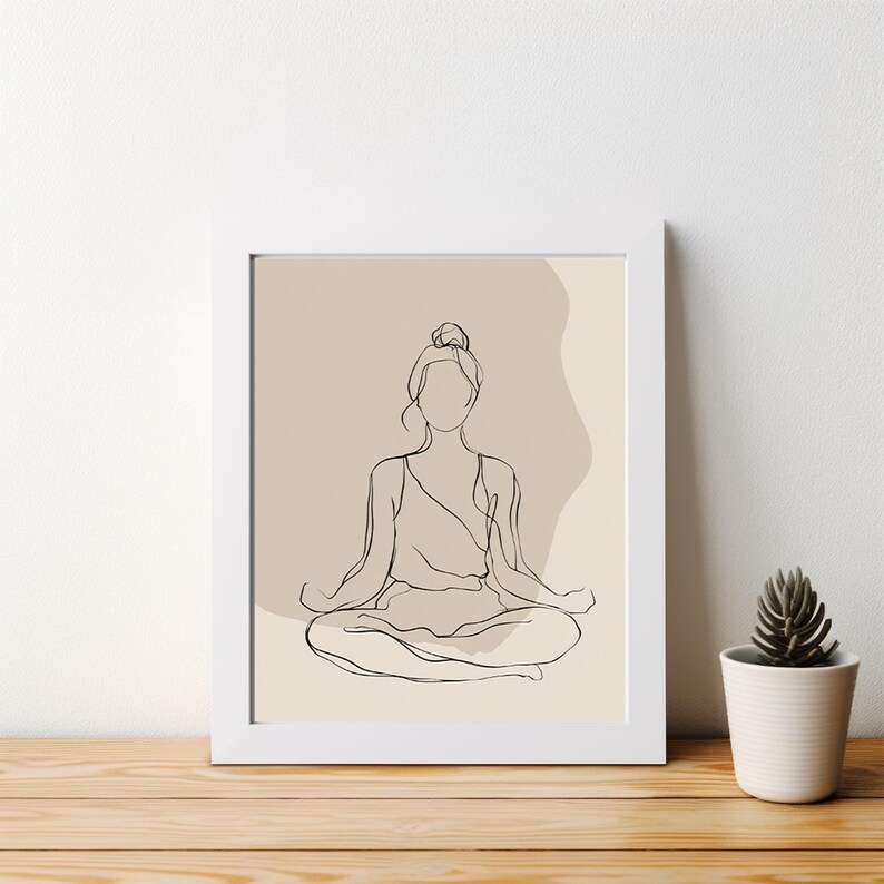 May include: A framed minimalist line art print depicting a person in a seated yoga pose. The artwork is in black against a beige background, framed in white. A small potted succulent sits on a wooden surface.