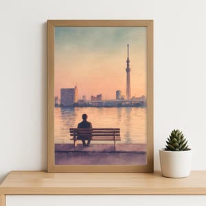 May include: Watercolor print of a person sitting on a bench overlooking a river and city skyline at sunset. The artwork is framed in a light brown wooden frame. A small potted succulent sits on a wooden shelf.