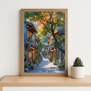 May include: A framed watercolor print of a Japanese street scene. The artwork depicts a cobblestone street lined with traditional buildings, lush greenery, and a large tree with orange and yellow leaves. The print is in a light brown wooden frame.