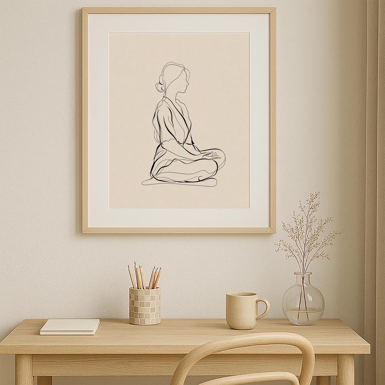 May include: A framed minimalist line art print. The artwork depicts a person in a seated meditation pose, rendered in black lines on a cream background. The frame is a light wood color. The print is displayed on a light wood desk with a mug, vase, and pencil holder.