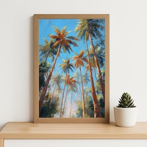 May include: Framed artwork depicting a view looking up at tall palm trees with orange and green fronds against a bright blue sky. The painting is in a wooden frame and sits on a light-colored wooden surface.