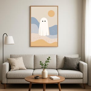 May include: A framed art print with a minimalist ghost design. The ghost is white with two black eyes, set against a backdrop of beige, blue, and yellow tones, with a sun graphic. The artwork is displayed above a grey sofa.