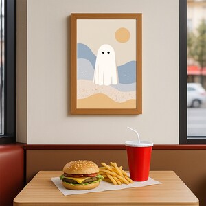 May include: A framed art print with a minimalist ghost illustration on a beach scene with a yellow sun. The image is displayed above a table with a burger, fries, and a red drink cup.