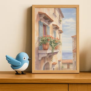 May include: A framed watercolour painting depicting a European building with a balcony adorned with flowers. The building features a light facade, red tiled roof, and blue shutters. A blue bird figurine is placed on a wooden surface in front of the painting.
