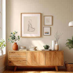 May include: A wooden cabinet with three framed art pieces hanging above it. The art includes a line drawing of a person, and two smaller framed pieces. A terracotta vase with greenery, books, and a green teapot are on the cabinet.