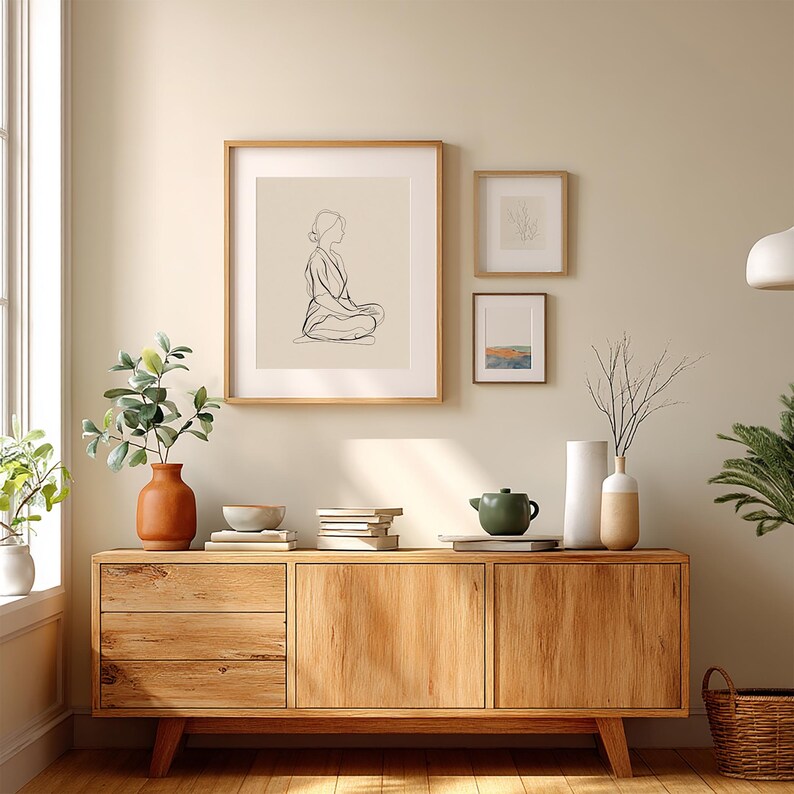 May include: A wooden cabinet with three framed art pieces hanging above it. The art includes a line drawing of a person, and two smaller framed pieces. A terracotta vase with greenery, books, and a green teapot are on the cabinet.