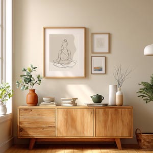 May include: A wooden sideboard with three framed artworks and decorative items. The largest frame displays a line drawing of a person in a seated yoga pose. A terracotta vase holds greenery, and a green teapot sits on the cabinet.