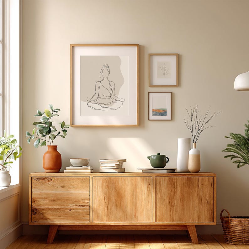 May include: A wooden sideboard with three framed artworks and decorative items. The largest frame displays a line drawing of a person in a seated yoga pose. A terracotta vase holds greenery, and a green teapot sits on the cabinet.