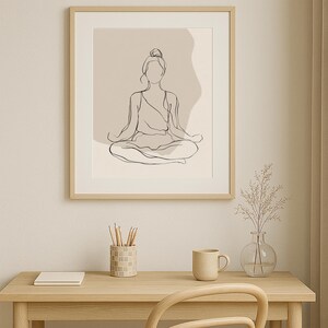 May include: A framed line art print of a person in a seated yoga pose. The artwork is minimalist, with a neutral colour palette. The frame is light wood. The print is displayed on a desk with a mug, pencils, and a vase.
