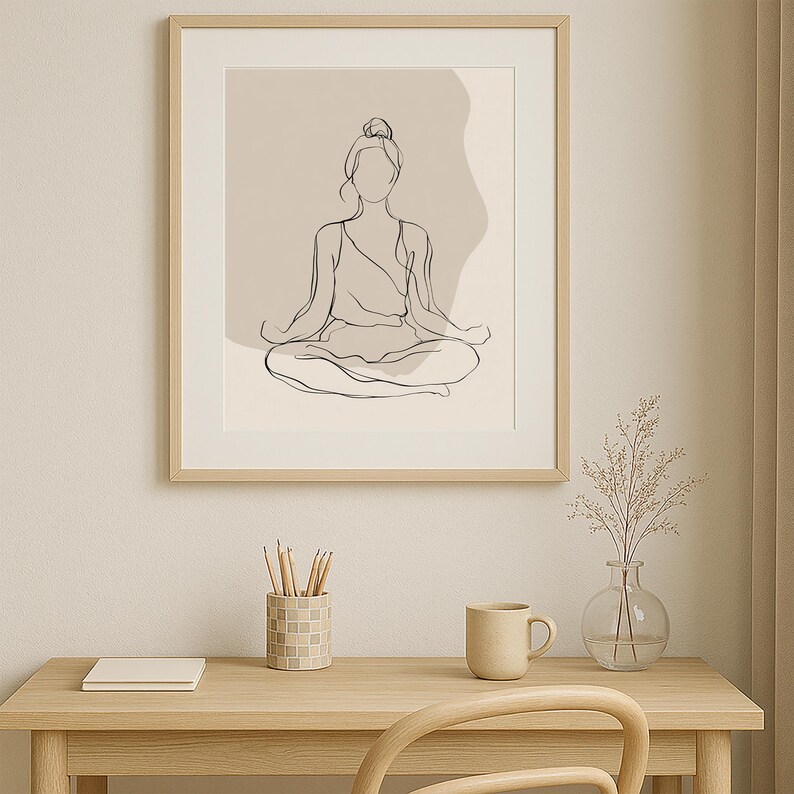 May include: A framed line art print of a person in a seated yoga pose. The artwork is minimalist, with a neutral colour palette. The frame is light wood. The print is displayed on a desk with a mug, pencils, and a vase.