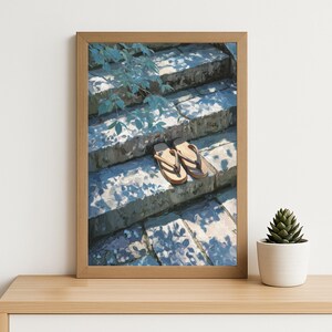 May include: A framed print of a pair of light brown flip-flops resting on a stone staircase. The stairs are shaded with leaf patterns. The artwork is in a wooden frame, with a small potted succulent plant on a wooden surface.