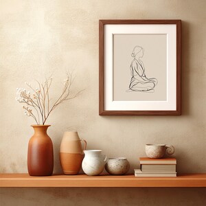 May include: A framed art print featuring a minimalist line drawing of a person in a seated meditation pose. The artwork is in a dark brown frame with a white mat. The image is displayed on a beige wall above a wooden shelf with vases and books.