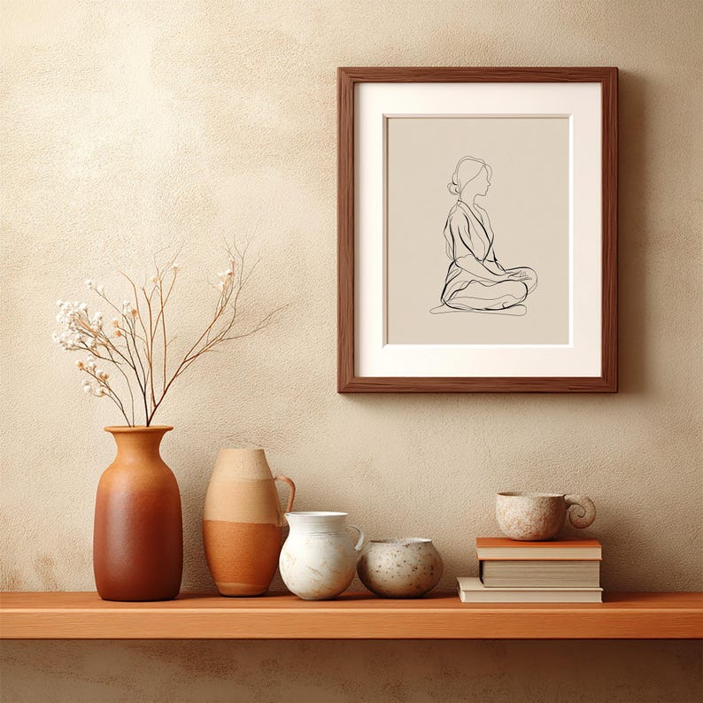 May include: A framed art print featuring a minimalist line drawing of a person in a seated meditation pose. The artwork is in a dark brown frame with a white mat. The image is displayed on a beige wall above a wooden shelf with vases and books.