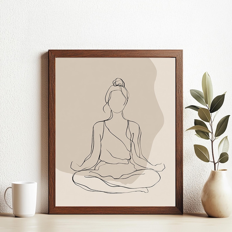 May include: A framed art print with a minimalist line drawing of a person in a seated yoga pose. The artwork is in black against a beige background, with a brown wooden frame. A white mug and a vase with greenery are in the foreground.