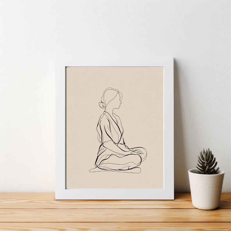 May include: A framed minimalist line art print. The artwork depicts a person in a seated meditation pose, rendered in black lines against a beige background. The white frame sits on a wooden surface, with a small potted plant to the right.