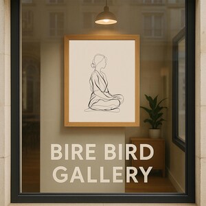 May include: A framed artwork featuring a minimalist line drawing of a seated figure in a meditative pose. The artwork is displayed in a window with the text "BIRE BIRD GALLERY" in large white letters. The frame is light brown.