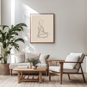May include: A framed art print featuring a minimalist line drawing of a person in a seated meditation pose. The artwork is in black against a beige background, set within a wooden frame. The print hangs on a neutral-colored wall in a living room setting.