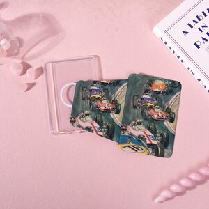 May include: Two coasters with a vintage race car design, featuring cars in various colors racing on a track. A clear plastic coaster holder and a book titled "A Table in Paris" are also present. The background is a soft pink.