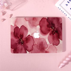 Peut inclure: Un autocollant pour ordinateur portable avec un motif floral de style aquarelle aux pétales bordeaux foncé et rose pâle. Le design présente une découpe centrale révélant le logo Apple blanc sur le capot, sur un fond rose clair.