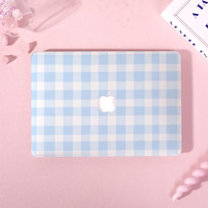 May include: A light blue and white gingham pattern laptop case with the Apple logo in the center. The case is on a pink surface, with a small glass container and a book visible in the background.