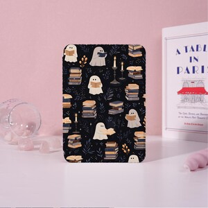 Op de afbeelding: Een zwart rechthoekig item met een herhalend patroon van witte spoken die boeken lezen, stapels boeken, kaarsen en bladeren. Het item is waarschijnlijk een notitieboek of een decoratief stuk. Een boek met de titel "A Table in Paris" is zichtbaar op de achtergrond.
