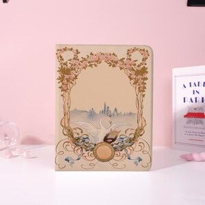 May include: A beige notebook with an Art Nouveau design featuring a swan, a cityscape, and floral elements. The cover has a decorative frame of pink roses and gold accents, with blue ribbons and a circular emblem.