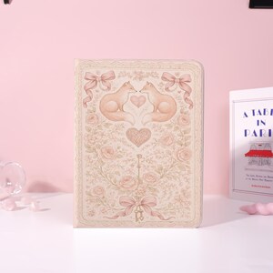 Könnte beinhalten: Ein dekoratives Notizbuch mit romantischem Design. Das Cover zeigt zwei Füchse, die sich gegenüberstehen, ein Herz, Rosen, Schleifen und einen Schlüssel. Die Farbpalette umfasst zarte Rosa-, Creme- und Beigetöne und erzeugt eine Vintage-Ästhetik.