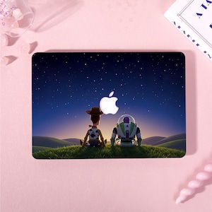 May include: A rectangular laptop skin featuring a graphic of two toy characters sitting on a grassy hill under a starry night sky. The design incorporates a white Apple logo in the center, positioned between the characters against the dark blue sky.