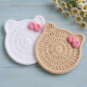 May include: Two circular crochet coasters shaped like cat heads, one in white and one in beige. Each coaster features a small, textured pink bow attached to the upper right ear area. The crochet work displays a consistent radial stitch pattern.