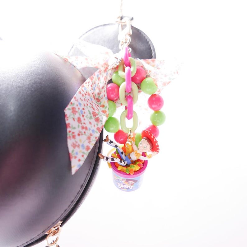 May include: A pink and green beaded charm with a small plastic figure of a cowboy riding a horse in a container of jelly beans. The charm is attached to a black purse with a floral ribbon.