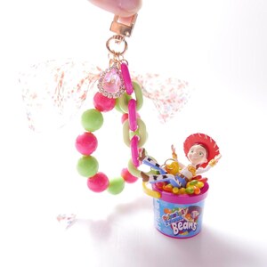 May include: A pink and green beaded charm bracelet with a pink and green chain attached to a pink and white jelly bean container with a cartoon character inside. The character is wearing a red hat and a blue shirt. The charm bracelet is attached to a pink and white bow. The text on the container reads "Jelly Belly Beans".