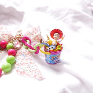 May include: A pink and green beaded chain with a charm of a toy cowboy character sitting in a colorful jelly bean container. The charm is attached to a pink and white floral ribbon.