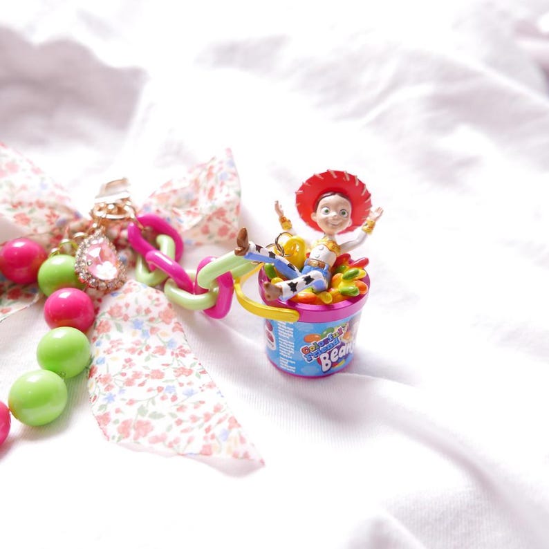 May include: A pink and green beaded chain with a charm of a toy cowboy character sitting in a colorful jelly bean container. The charm is attached to a pink and white floral ribbon.