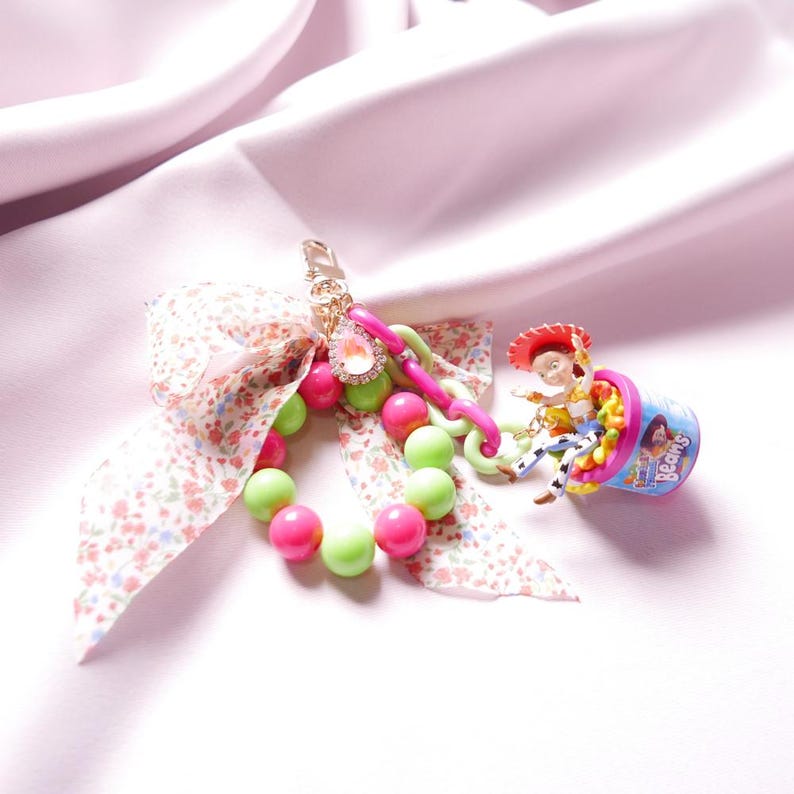 May include: A pink and green beaded keychain with a floral ribbon and a toy character riding in a container of jelly beans. The container has the word "Beans" on it.