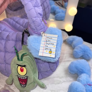 May include: A quilted lavender handbag with a matching handle and a cartoon character plushie attached. A blue fluffy heart-shaped charm with a handwritten note that says "Mom I'm trying to smile" is also attached.