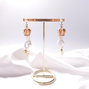 Op de afbeelding: Een paar gouden en witte oorhangers met een hand die een kleine kroon vasthoudt. De kroon is versierd met witte parels.