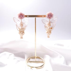 Può includere: Un paio di orecchini dorati con una statuetta di cherubino, una rosa rosa e ali di farfalla trasparenti.