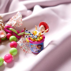 May include: A pink and green beaded bracelet with a charm of a toy cowboy doll sitting in a pink and blue container of jelly beans. The container has the text "Colorful Friend Beans" on it.