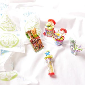 May include: Toy Story themed miniature figurines and snacks.  A green alien in a red and white popcorn container, a green alien in a clear tube with yellow candy, a green alien in a glass jar, a cowboy in a red and white bean container, and a cowboy in a green and yellow pizza sticks box.