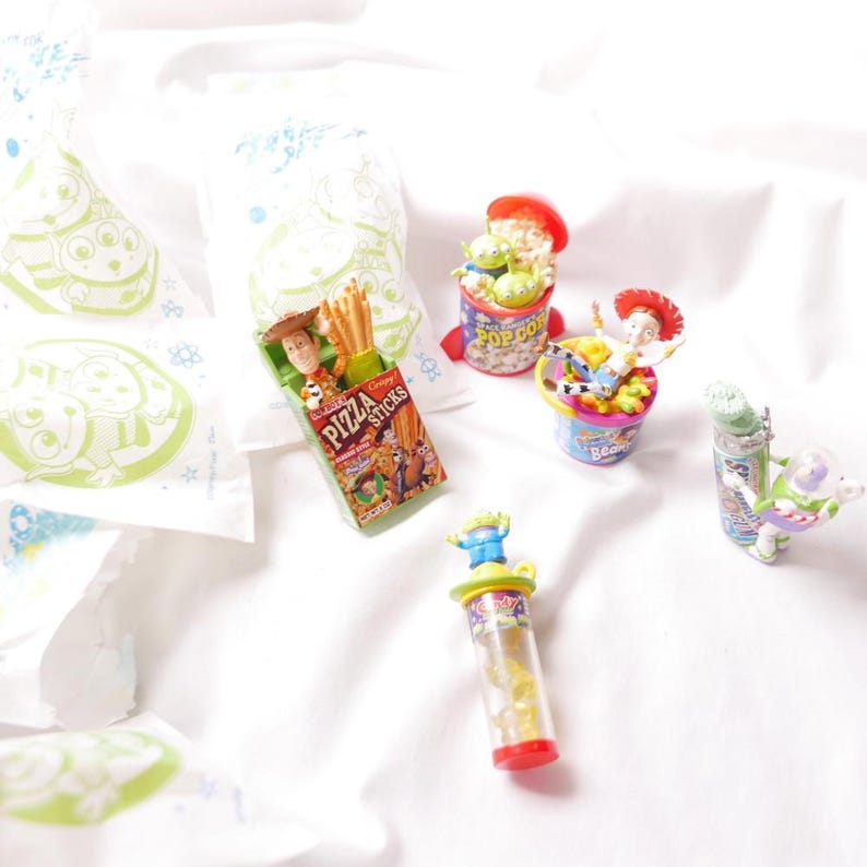 May include: Toy Story themed miniature figurines and snacks.  A green alien in a red and white popcorn container, a green alien in a clear tube with yellow candy, a green alien in a glass jar, a cowboy in a red and white bean container, and a cowboy in a green and yellow pizza sticks box.