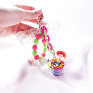 May include: A pink and green beaded bracelet with a charm of a toy story character, Woody, sitting in a bucket of jelly beans. The charm is attached to a gold chain with a pink and green plastic chain. The bracelet is tied with a white ribbon with pink flowers.