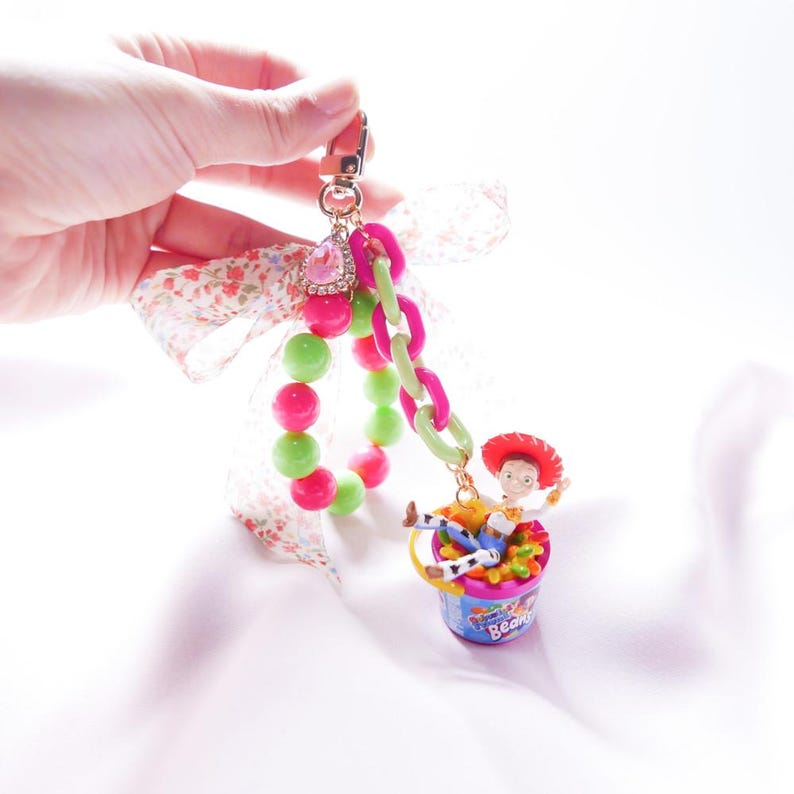 May include: A pink and green beaded bracelet with a charm of a toy story character, Woody, sitting in a bucket of jelly beans. The charm is attached to a gold chain with a pink and green plastic chain. The bracelet is tied with a white ribbon with pink flowers.