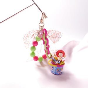 May include: A pink and green beaded charm with a pink and green chain, a pink and white floral ribbon, and a small plastic toy figure of Woody from Toy Story sitting in a blue and pink bucket with the words "Rainbow Beach" on it.
