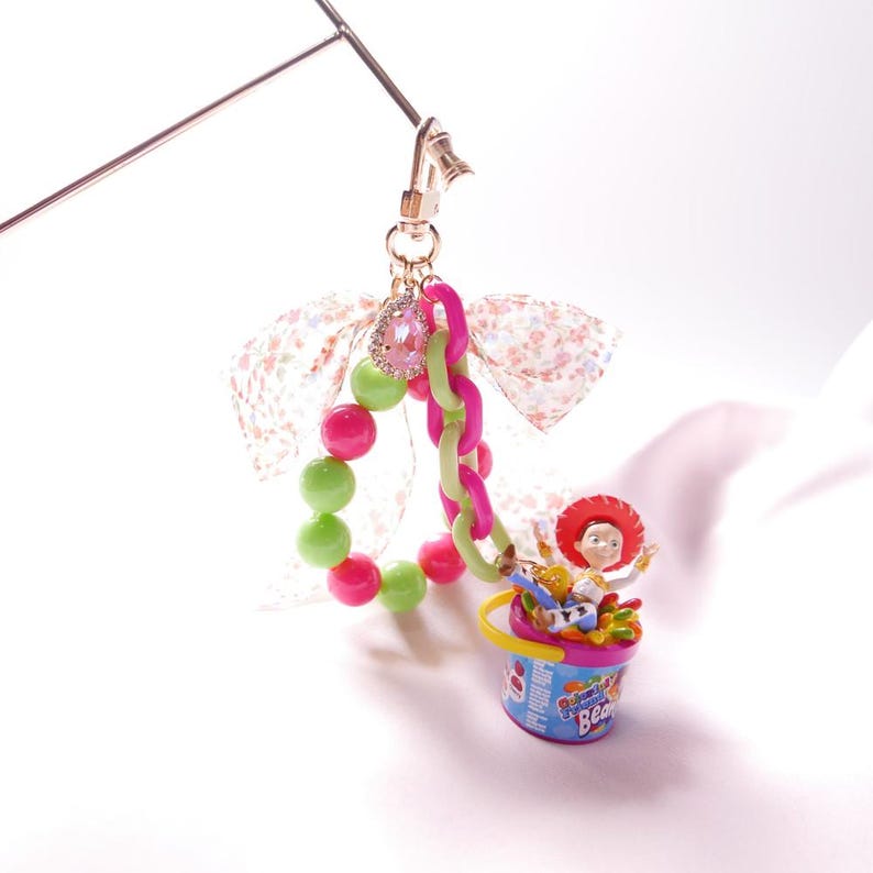 May include: A pink and green beaded charm with a pink and green chain, a pink and white floral ribbon, and a small plastic toy figure of Woody from Toy Story sitting in a blue and pink bucket with the words "Rainbow Beach" on it.