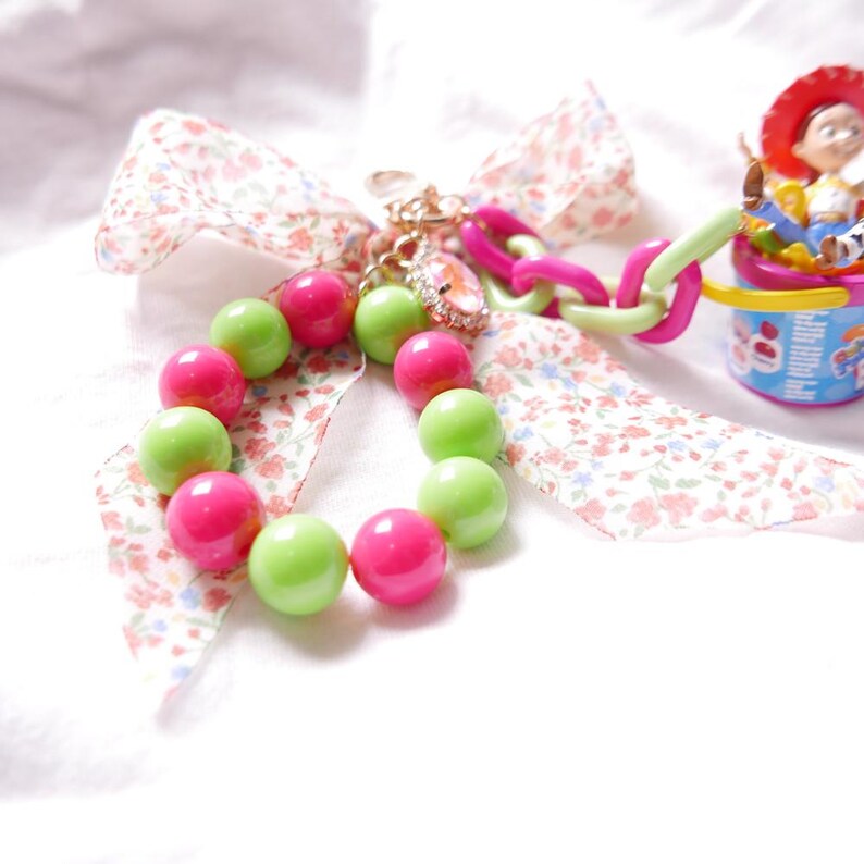 May include: A pink and green beaded charm bracelet with a floral ribbon and a toy character keychain. The keychain features a cowboy character sitting in a bucket.