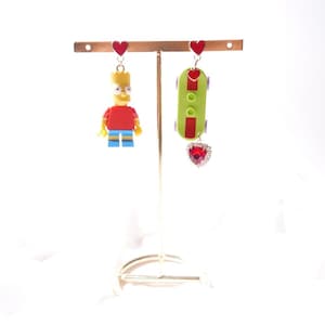 May include: Novelty earrings on a gold stand. One earring is a Bart Simpson figure, the other a green skateboard with a red heart gem. Both have red heart studs. The earrings are displayed on a gold-coloured stand.
