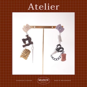 May include: Earrings with sewing-themed charms. One earring features a sewing machine, measuring tape, and needle. The other has scissors, a thimble, and thread. The word "Atelier" is at the top. The earrings are suspended from a gold-colored bar.